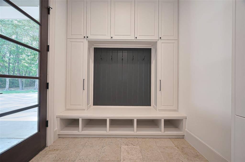 Bright and clean mudroom with built-in storage. It features white cabinetry, multiple hooks for hanging, and cubbies below for shoes or accessories. The large, windowed door allows natural light to fill the space and easy access to outside.