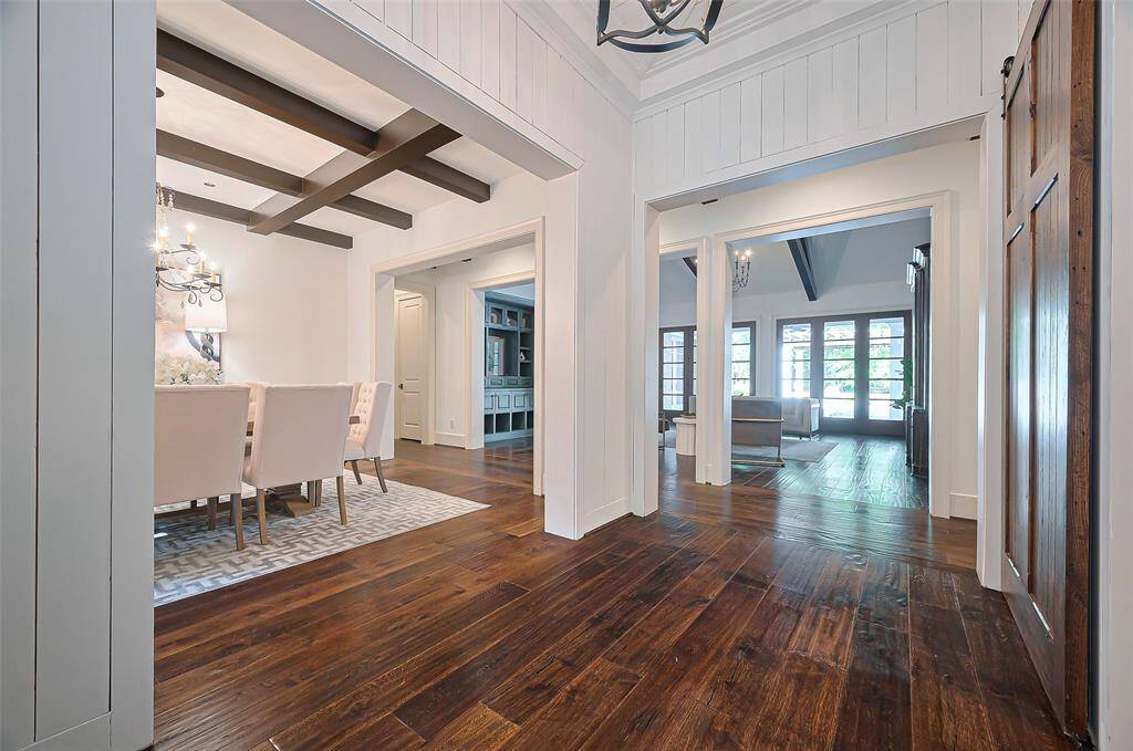 Bright, open hallway with rich hardwood floors, leading to a dining area with a stylish chandelier and a spacious living room featuring large windows and exposed beams.