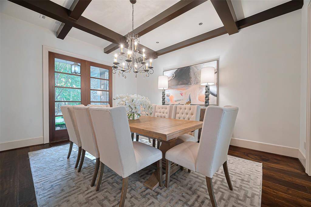 Elegant dining room features a modern chandelier, and a stylish ceiling with exposed beams. Large windows provide natural light, complemented by tasteful artwork and a neutral color palette, creating a welcoming and sophisticated atmosphere.