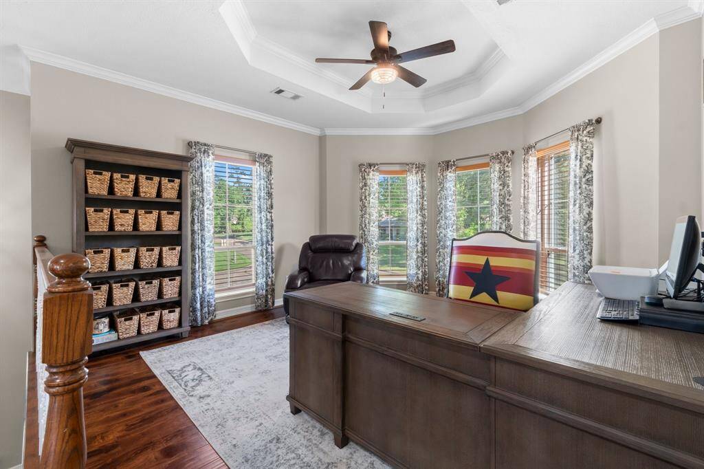 Bright, well-appointed home office with a large window bay, providing ample natural light. The room features a ceiling fan, elegant desk, comfortable chair, and shelving with organized storage baskets, offering both functionality and style.