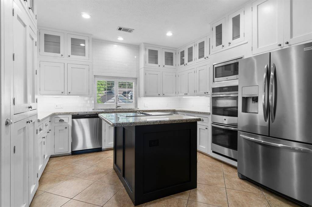 This modern kitchen features sleek stainless steel appliances, abundant white cabinetry with glass accents, and a central island with a dark finish. It boasts a bright, open feel with recessed lighting and a window above the sink.