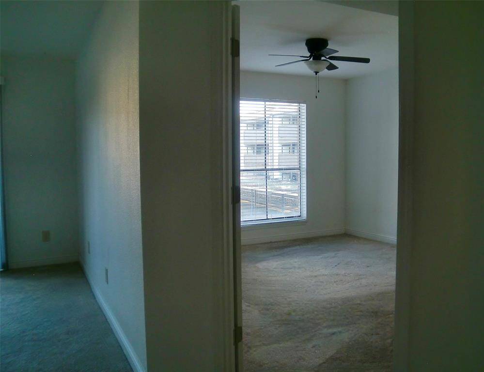 This photo shows a carpeted room with neutral walls. It features a window with blinds and a ceiling fan, providing natural light and ventilation.