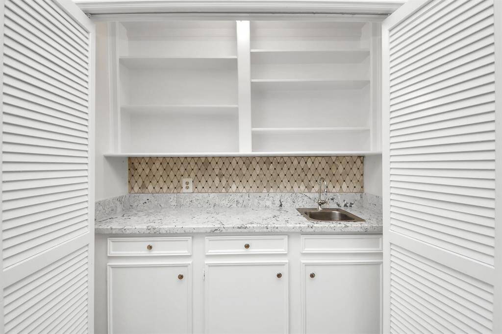 This photo shows a compact wet bar or pantry area with white cabinetry, open shelving, and a small sink. It features a granite countertop and a decorative tile backsplash, enclosed by white louvered doors.