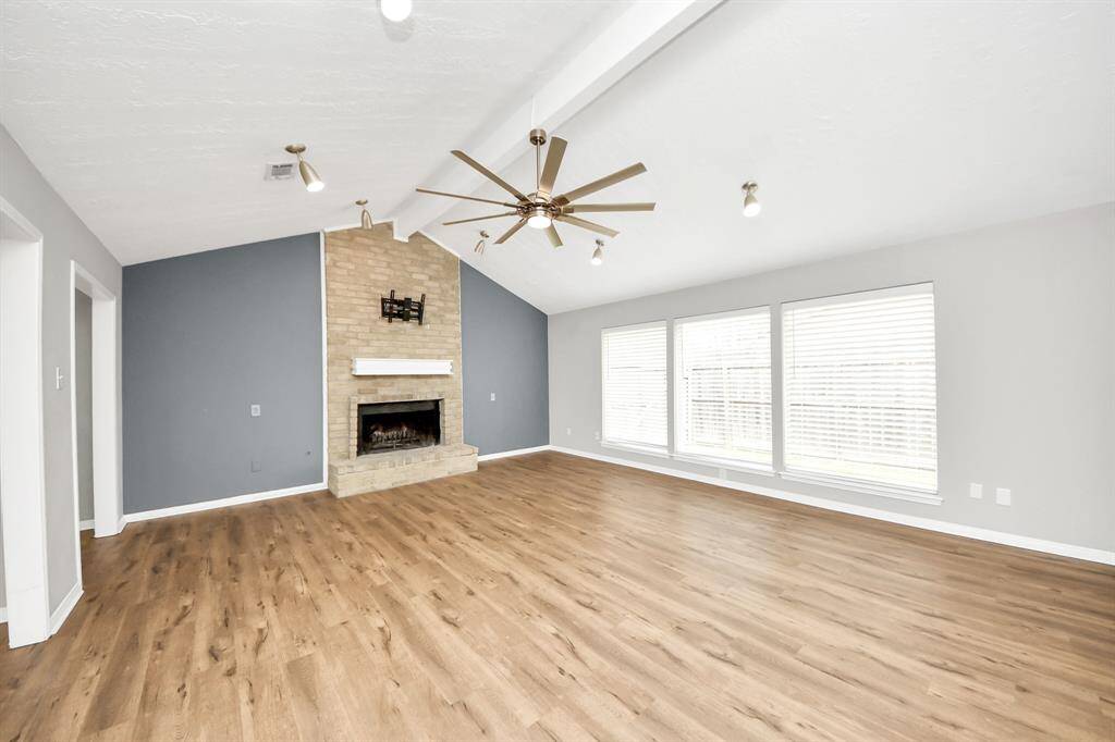 This spacious living room features a vaulted ceiling with a modern ceiling fan, a cozy brick fireplace, and abundant natural light from large windows. The wood flooring adds warmth, while the neutral wall colors provide a versatile backdrop.