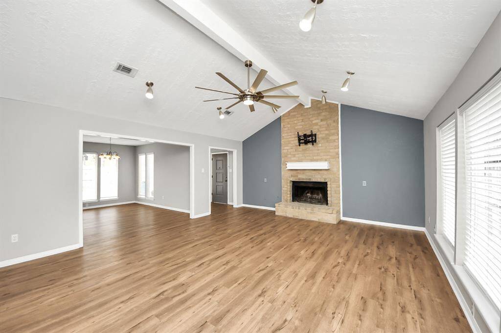 This spacious living area features a vaulted ceiling with a modern fan, a cozy brick fireplace, and ample natural light from large windows. The room's open layout and wood flooring create a warm, inviting atmosphere, ideal for entertaining or relaxing.