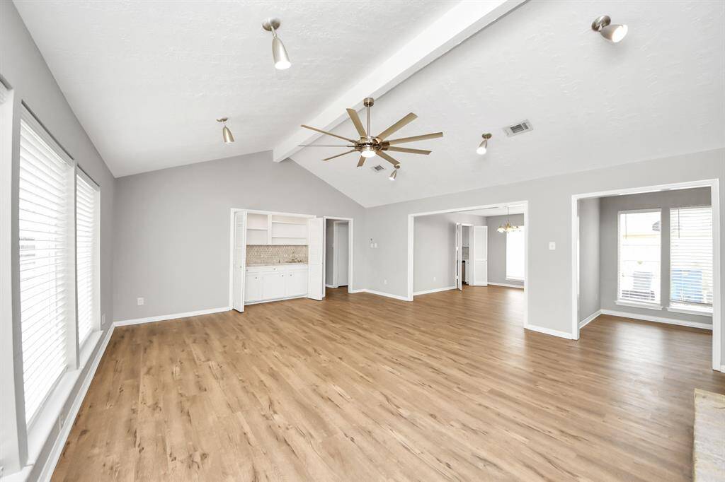 This spacious living area features vaulted ceilings with modern lighting, wood-look flooring, and large windows for ample natural light. It includes built-in shelving and connects seamlessly to dining spaces, creating an open and airy feel.