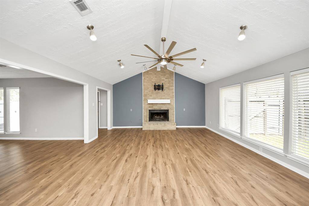 This spacious living room features a vaulted ceiling with a modern fan, a brick fireplace, and ample natural light from large windows with blinds. The wood-look flooring adds warmth, and the neutral walls offer a fresh, inviting atmosphere.