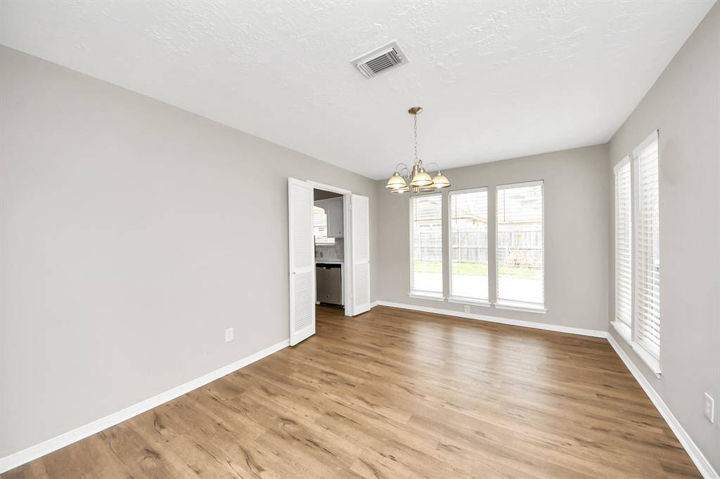 Bright dining area with large windows, wood flooring, and neutral walls. Features a central light fixture and has direct access to the kitchen through a doorway.