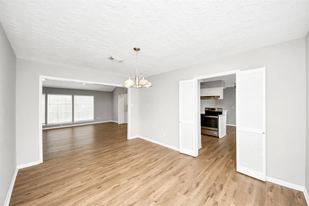 This photo showcases a spacious, open-concept living area with light wood flooring. It features a dining area with a central light fixture, leading into a kitchen with white cabinetry and stainless steel appliances. Large windows in the adjacent living room provide plenty of natural light. The walls are painted in a neutral gray color, creating a modern and inviting atmosphere.