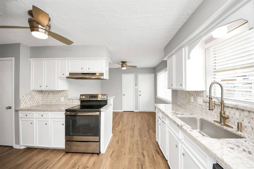 Bright, modern kitchen with white cabinetry, light granite countertops, and stainless steel appliances. It features wood flooring, a stylish backsplash, and ample natural light through large windows. Ceiling fans add a contemporary touch.