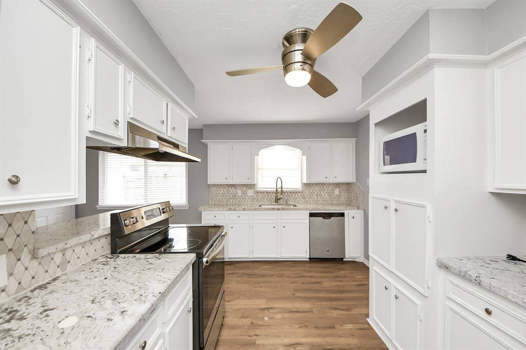 Modern kitchen with white cabinetry, granite countertops, and stainless steel appliances. Features a stylish ceiling fan and wood flooring. Bright and spacious with ample storage and a built-in microwave.