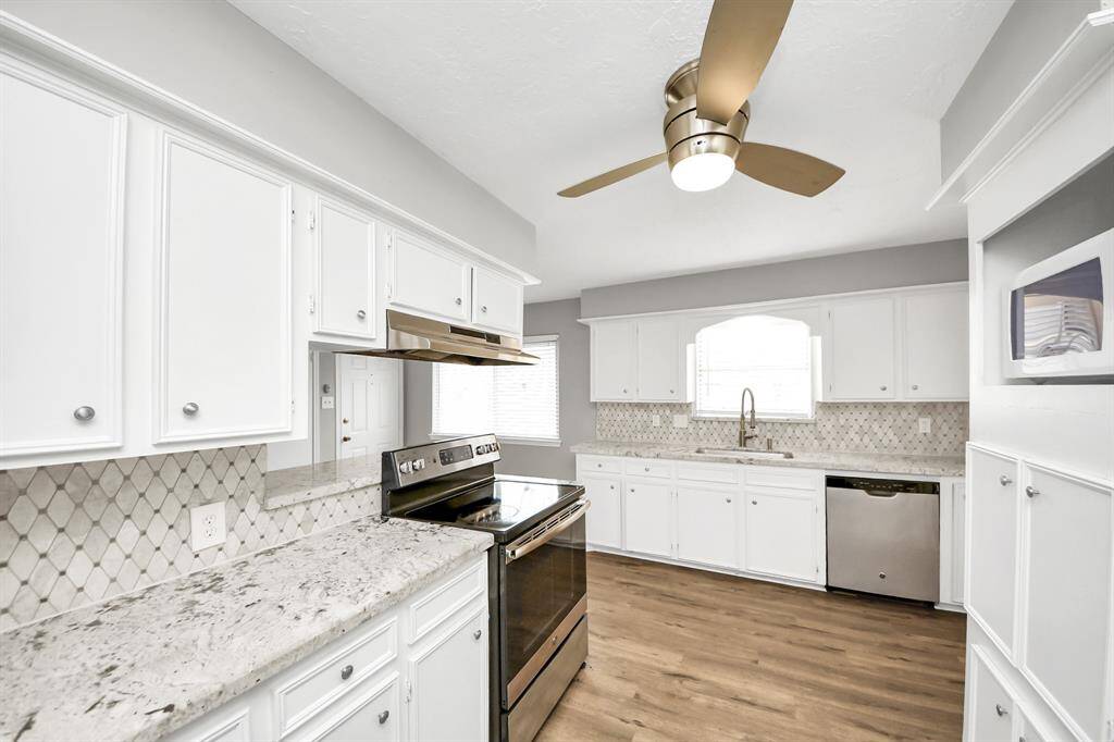 This kitchen features modern appliances, white cabinetry, and a sleek marble-look countertop. The backsplash adds texture, while the ceiling fan offers a contemporary touch. Natural light streams through a window above the sink, enhancing the bright and airy feel.