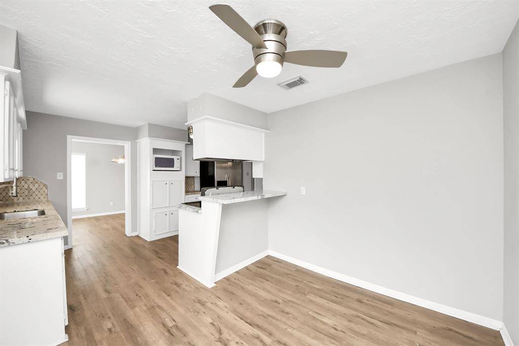 This photo showcases a modern kitchen and dining area with light gray walls and wood flooring. The kitchen features white cabinetry, granite countertops, and a built-in microwave. A ceiling fan adds comfort to the space. The open layout leads to a well-lit adjoining room.