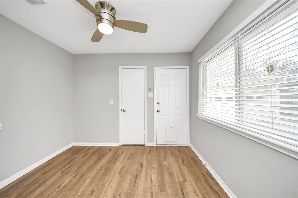 This room features light gray walls and wood flooring, with a large window allowing natural light to fill the space. A ceiling fan adds comfort, and there are two doors, one possibly leading outside.