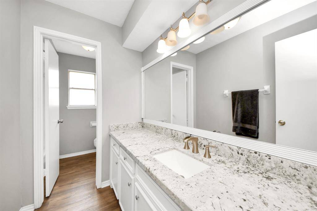 This bathroom features a long granite countertop with a sleek under-mount sink and modern fixtures. It has ample cabinet storage, a large mirror with stylish lighting, and a neutral color palette. The space is bright and airy, enhanced by natural light and warm wood flooring.