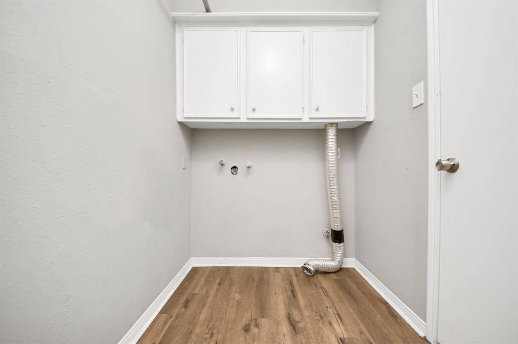 This is a compact laundry room with light gray walls and wood flooring. It features upper white cabinets for storage and hookups for a washer and dryer. An exposed dryer vent is visible on the right.