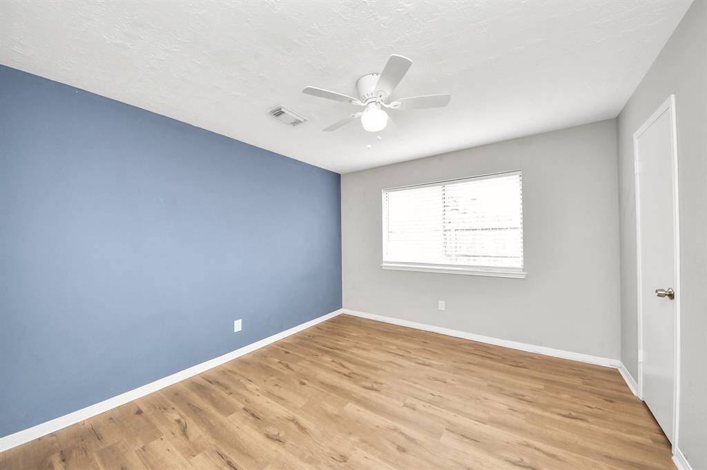 This room features a modern design with a blue accent wall, light gray walls, and wood-style flooring. A large window provides natural light, and a ceiling fan adds comfort. Ideal for a bedroom or office space.