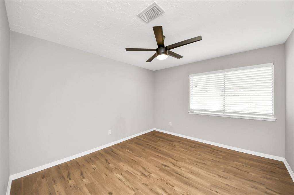 This room features fresh gray walls, wood-style flooring, a modern ceiling fan, and ample natural light from a large window with blinds.
