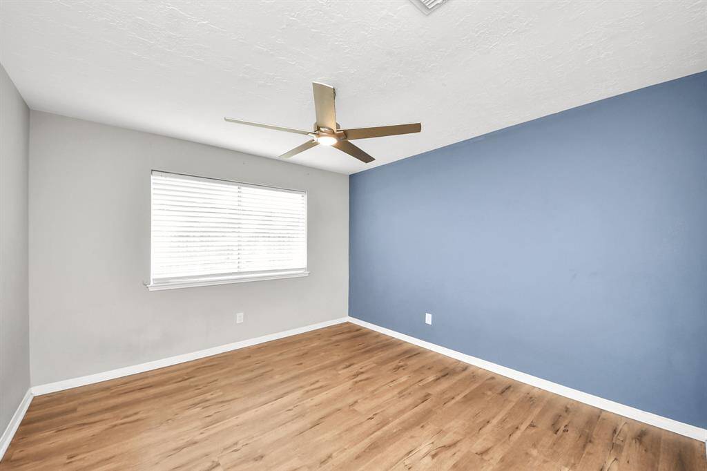 This room features a modern ceiling fan, wood-look flooring, a light gray wall with a large window, and an accent blue wall, creating a bright and inviting space.