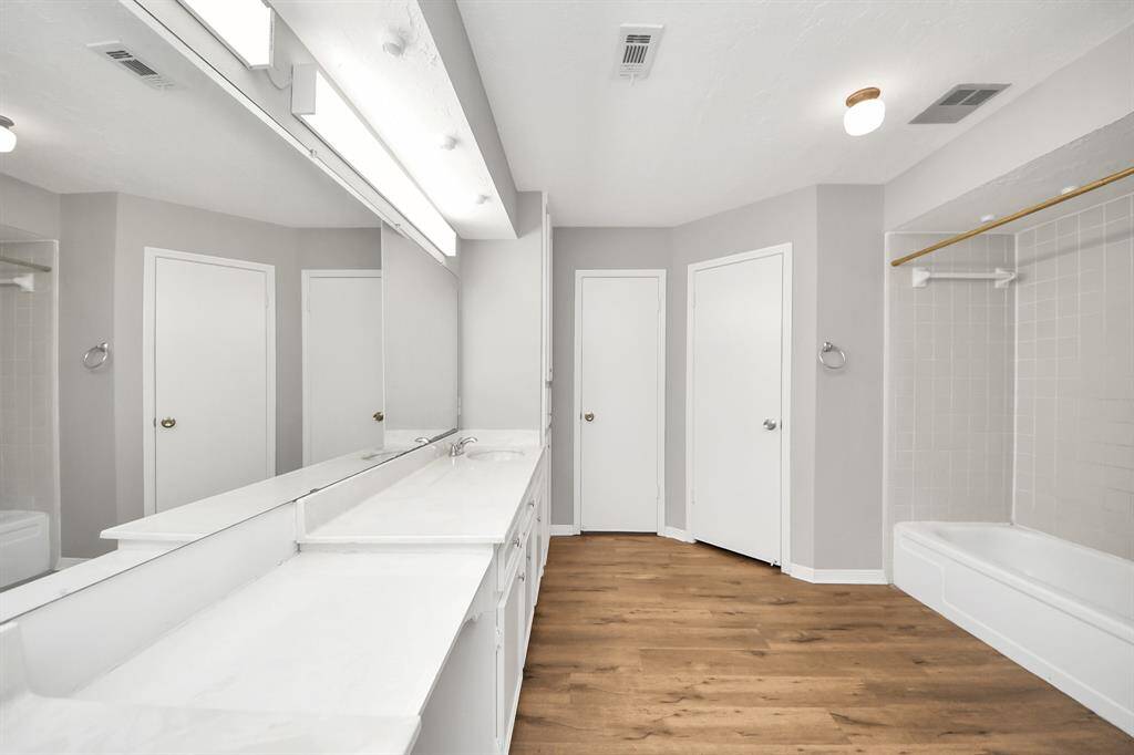 This bathroom features a spacious layout with a long vanity and dual sinks, ample counter space, a large mirror, and a combination shower and bathtub. The room is well-lit and has wood-style flooring for a modern touch.