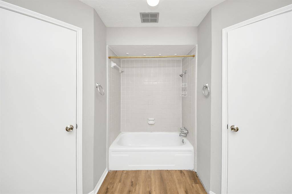 This bathroom features a clean, minimal design with neutral walls, a white bathtub with a tiled shower area, and wood-style flooring. Two doors on either side provide access, and there are towel rings for convenience.