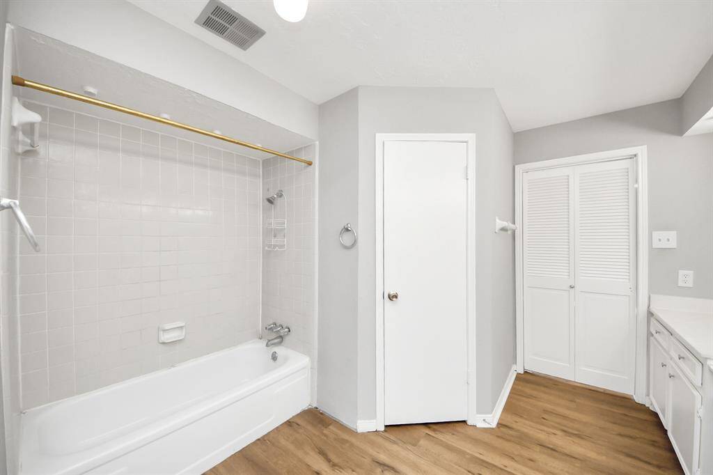 This bathroom features a clean design with a white bathtub and tiled shower area, complemented by light gray walls. It has wood-look flooring, a storage closet with louvered doors, and a simple vanity for a fresh, modern look.