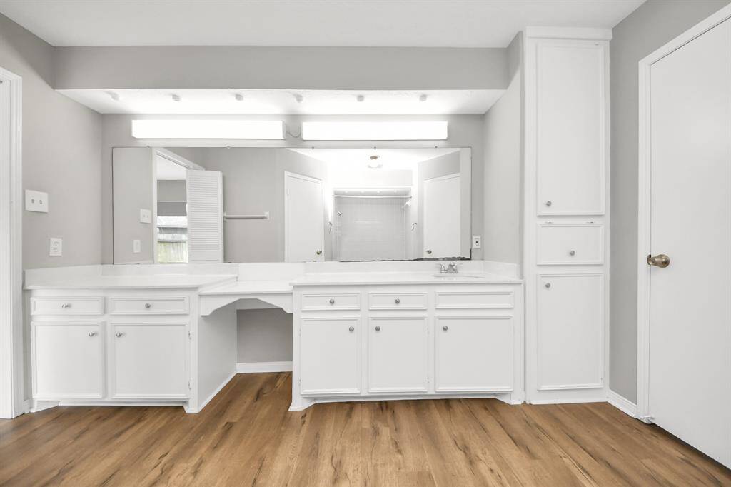 This photo showcases a spacious bathroom with white cabinetry and plenty of storage. It features a large mirror and bright lighting, complemented by a wood-style floor and neutral walls, creating a clean and modern look.