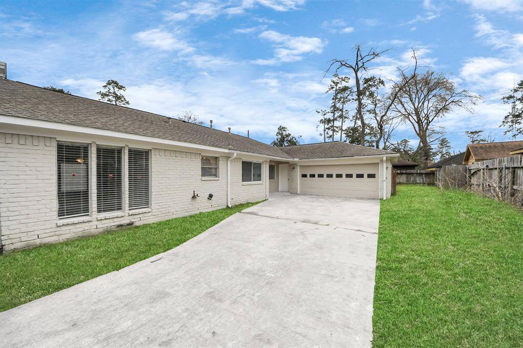 This photo features a single-story home with a spacious driveway leading to a two-car garage. The exterior is painted brick, and the house has a well-maintained front lawn. Large windows offer plenty of natural light, and the property is fenced for privacy.