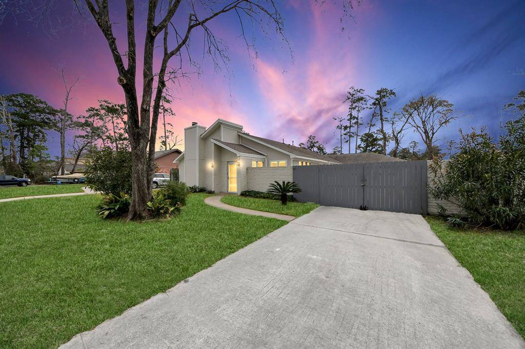 This photo showcases a modern home with a sleek design, featuring a well-maintained lawn and a long driveway leading to a wooden gate. The sky is a stunning blend of pink and purple hues, adding a serene backdrop to the property.