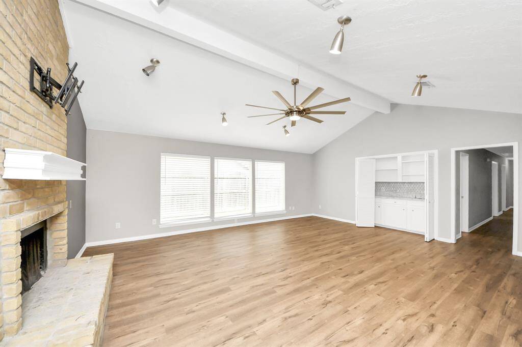 Bright, spacious living room with vaulted ceiling, modern light fixtures, and wood flooring. It features a brick fireplace, large windows, built-in shelves, and a ceiling fan.