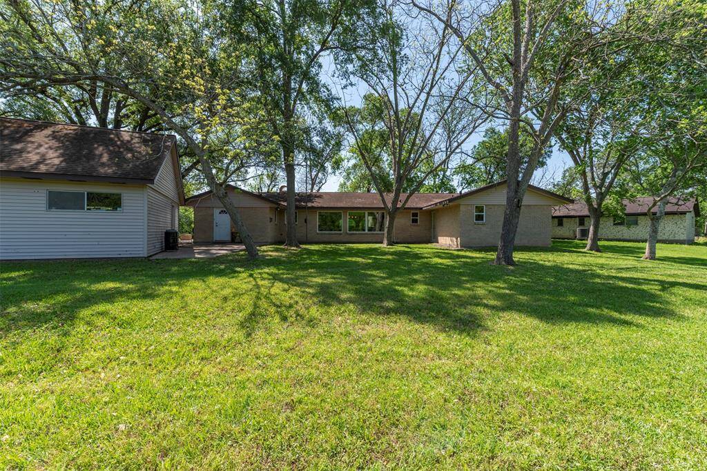 This view from the spacious backyard shows the full rear exterior of the home, framed by trees and wide open green space. Large windows line the back wall, offering peaceful views. The detached garage and shaded patio area create a versatile set up for out door living or entertaining with plenty of room for a pool.