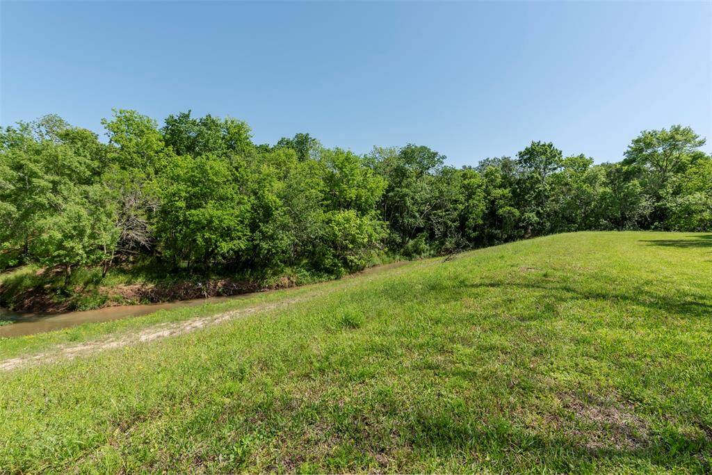 A grassy sloped side lot reveals gentle rolling terrain leading to Clear Creek with  thick tree coverage along the water's edge and open green space in the foreground.