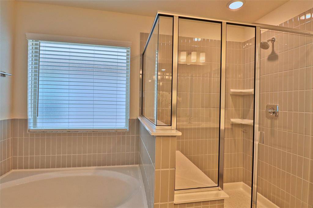 This modern primary bathroom features a large window with blinds, a spacious bathtub, and a glass-enclosed shower with sleek fixtures, offering a bright and inviting space.
