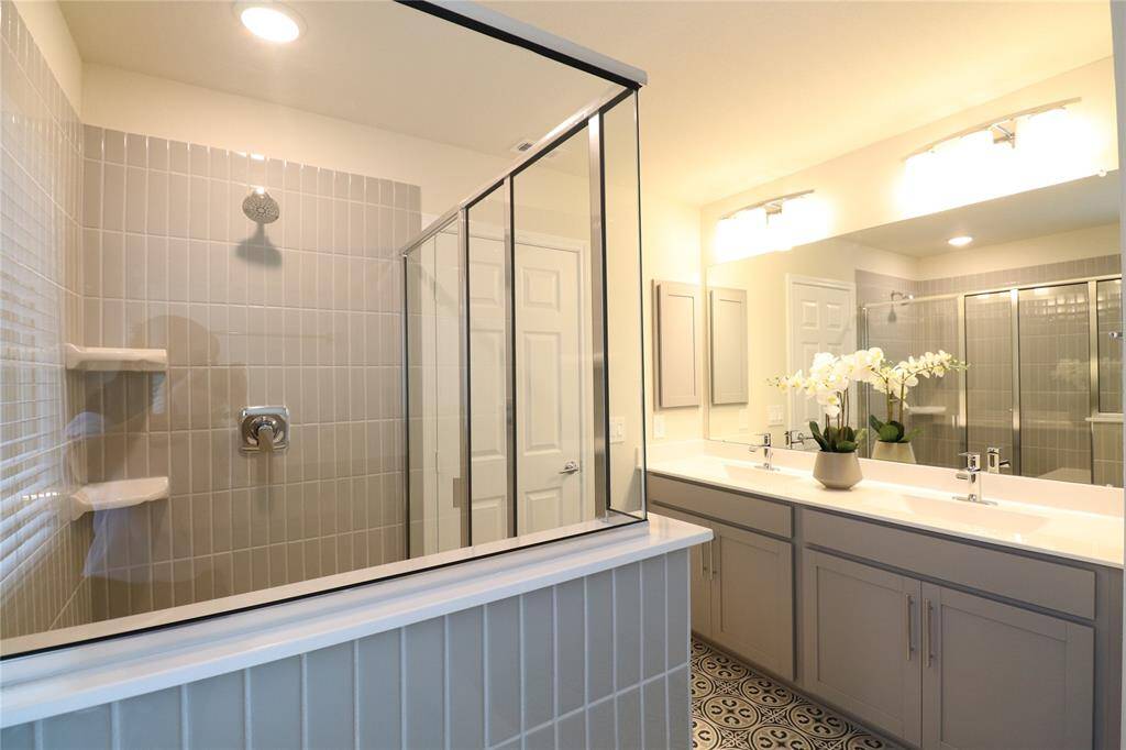 Bright and modern bathroom featuring a glass-enclosed shower, dual sinks with a large mirror, stylish cabinetry, and decorative tile flooring.