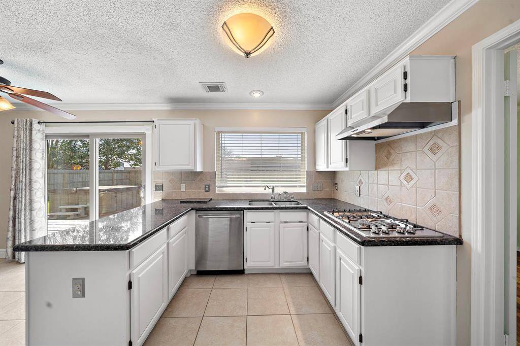 This kitchen features white cabinetry, a black granite countertop, and a modern gas range. It has a tile backsplash and a window above the sink for natural light looking outside to your backyard perfect for grillin & chillin!