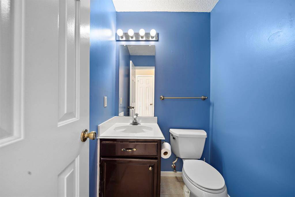 This bathroom features vibrant blue walls, a vanity with a sink, a mirror with overhead lighting, and a toilet. The space is compact yet functional, with a towel bar and neutral tile flooring.