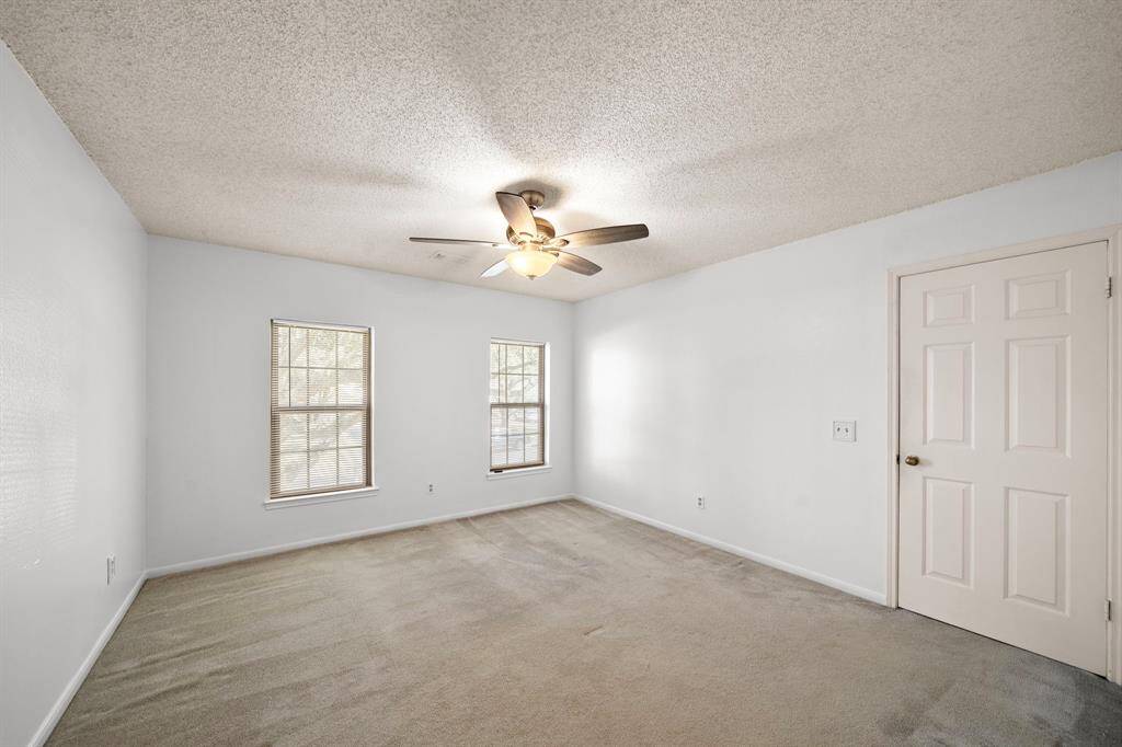 This spacious bedroom features neutral carpeting, a ceiling fan, and ample natural light from two large windows. The light-colored walls offer a versatile backdrop for any decor style.