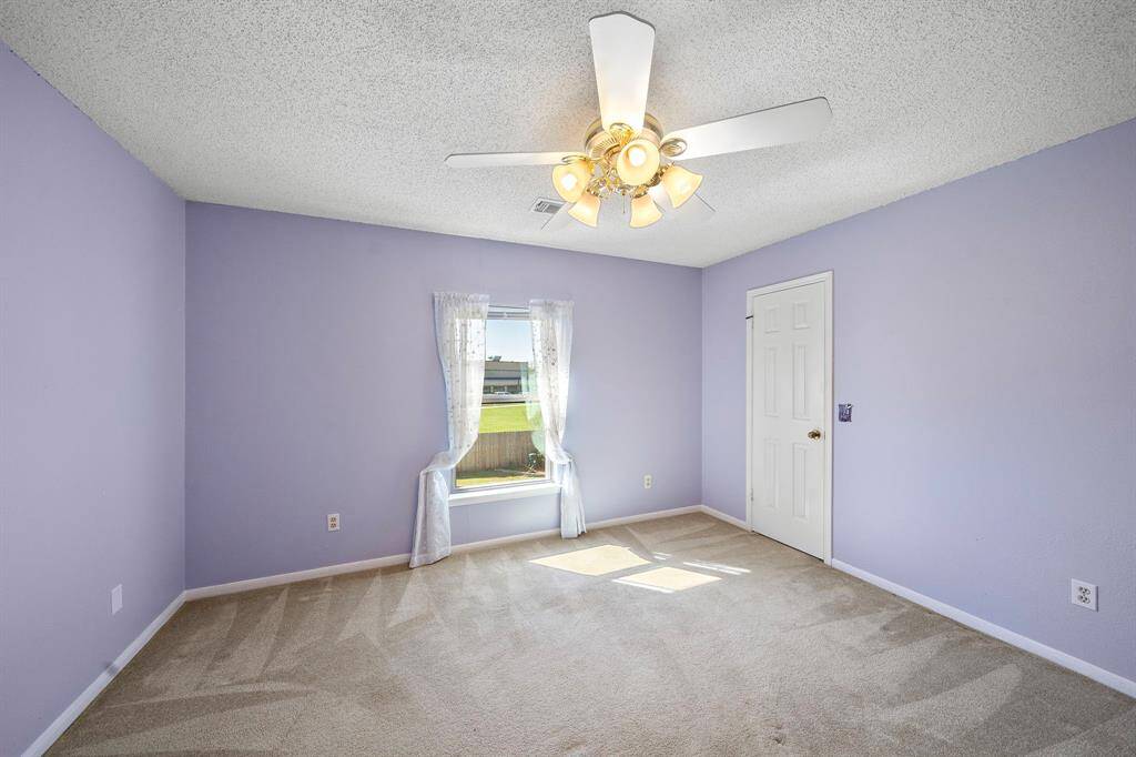 This bedroom features soft lavender walls, a ceiling fan with lights, and a carpeted floor. A window with sheer curtains allows natural light to brighten the space, and there's a door for additional storage.