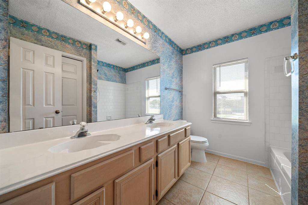 This bathroom features a dual-sink vanity with light wood cabinets, a large mirror with ample lighting, and a bathtub with shower. It has natural light from a window and a decorative blue and seashell-themed wallpaper border.