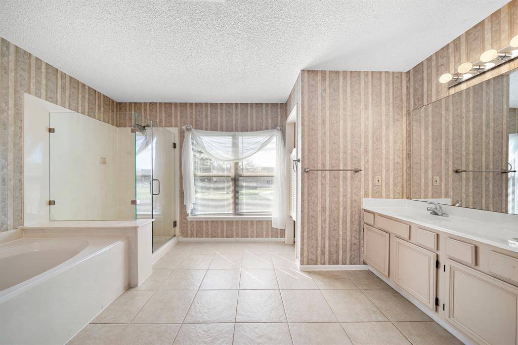 This spacious bathroom features a large soaking tub, separate glass-enclosed shower, and a double vanity with ample storage. The room is brightened by a window with sheer curtains, and the walls are adorned with subtle striped wallpaper.