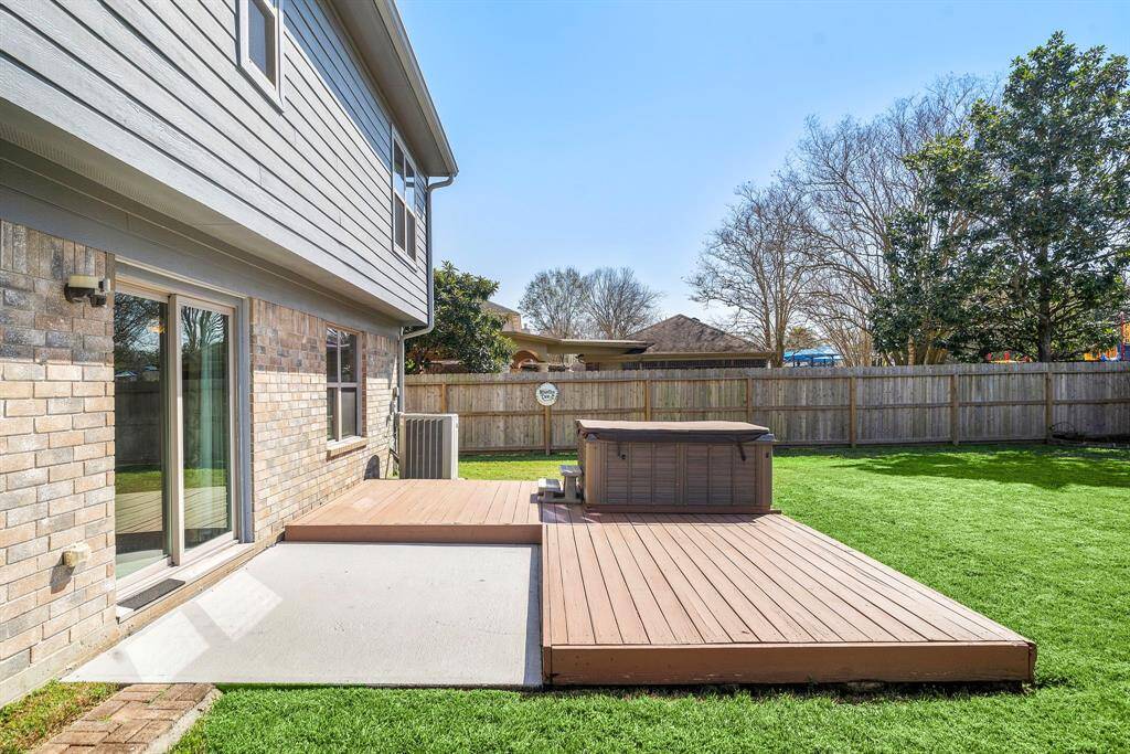 This photo shows a well-maintained backyard with a spacious deck area and a hot tub. The yard is fenced, providing privacy, and features a lush green lawn. The house exterior has a modern look with sliding glass doors leading to the patio. Ideal for outdoor relaxation and entertainment.