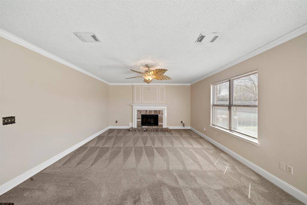 Spacious living room with neutral-toned walls, featuring a cozy fireplace and large window for natural light. Carpeted flooring and a ceiling fan add comfort and style.