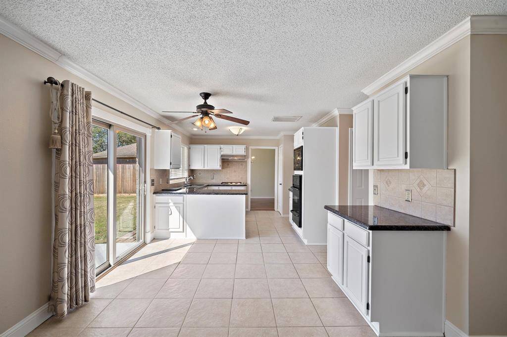 This kitchen features bright white cabinetry, granite countertops, and a tile backsplash. It has a ceiling fan, tiled flooring, and sliding glass doors that open to a backyard, providing ample natural light.