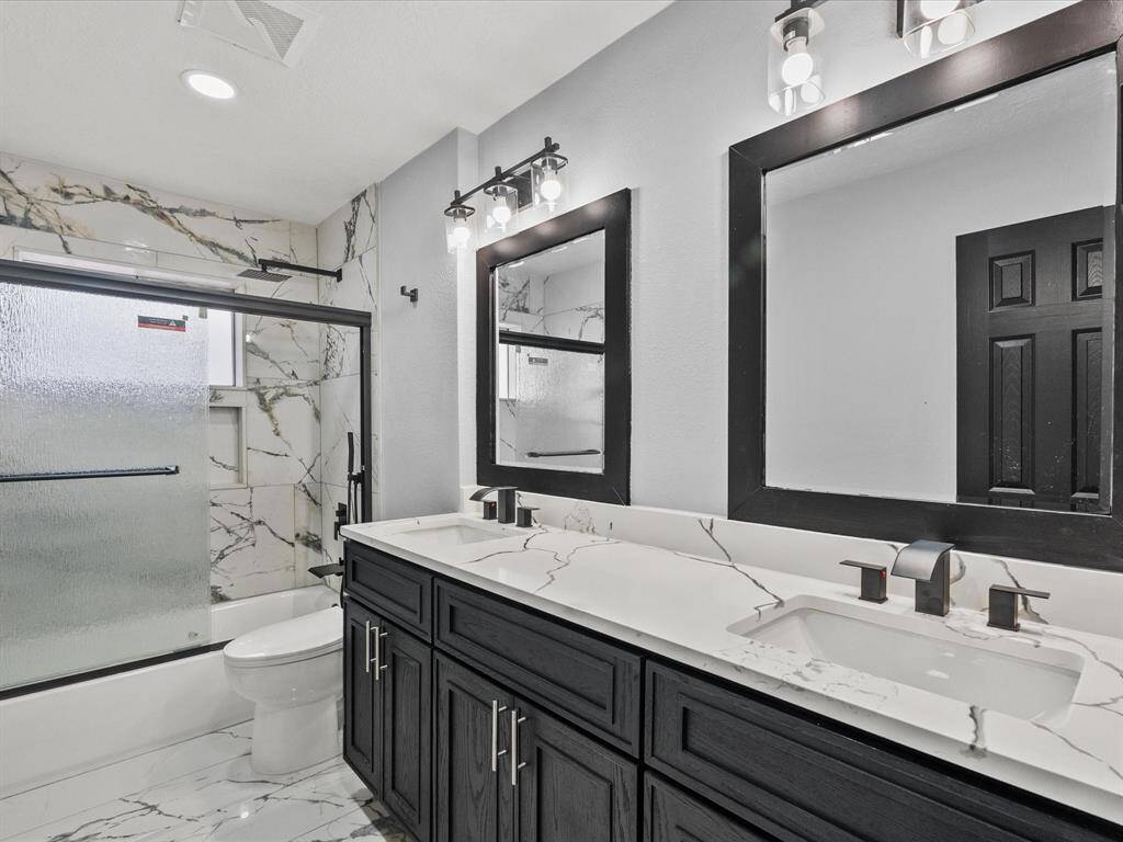 This bathroom features a modern design with porcelain tiles, a double vanity with sleek black cabinetry, and matching framed mirrors. It includes a bathtub with a frosted glass sliding door, contemporary lighting, and a clean, cohesive aesthetic.