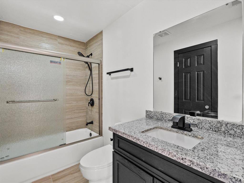 Modern bathroom with a sleek design, featuring a granite countertop and a black vanity. It includes a bathtub with a glass shower door and wood-look tile walls, complemented by black fixtures and a black door for a contemporary feel.