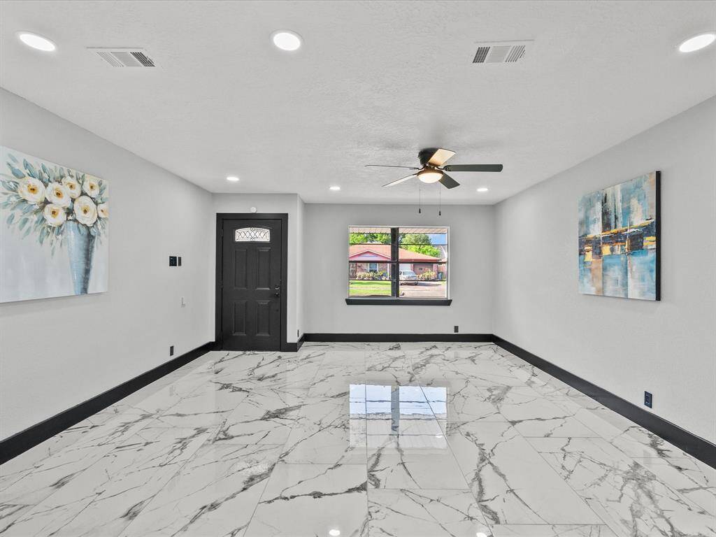 This living room features a modern design with sleek porcelain flooring neutral walls, and recessed lighting. A ceiling fan is centrally placed, and large windows allow for natural light. The space is accented with contemporary artwork and a dark front door.