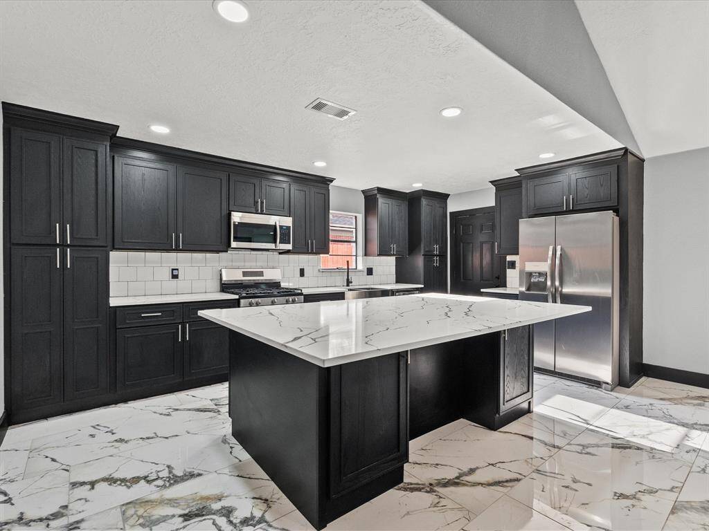 This modern kitchen features sleek black cabinetry, stainless steel appliances, and a spacious quartz-patterned island. The white subway tile backsplash and porcelain tile flooring enhance the clean, contemporary look. Ideal for cooking and entertaining.