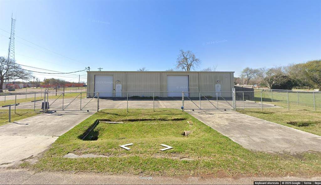 This photo shows a large, single-story metal building with two garage doors, set behind a chain-link fence. The property includes a paved driveway and some grassy areas, situated on a spacious lot.