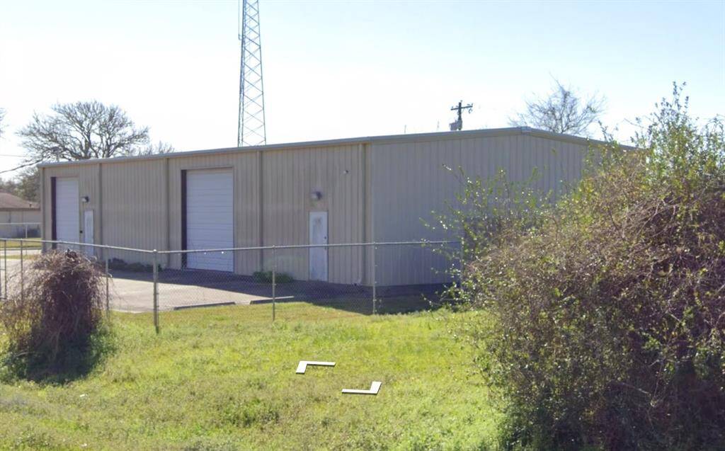 This photo shows a large metal warehouse or garage with two roll-up doors and a side entrance. It's surrounded by a chain-link fence and situated on a grassy lot, ideal for storage or light industrial use.