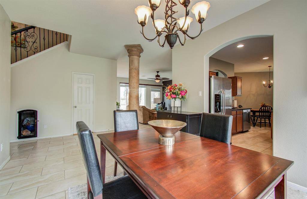 This inviting dining area features an elegant chandelier, a wooden table with leather chairs, and a view into the kitchen with stainless steel appliances. An open layout connects the living space, accentuated by a decorative column and neutral tile flooring.